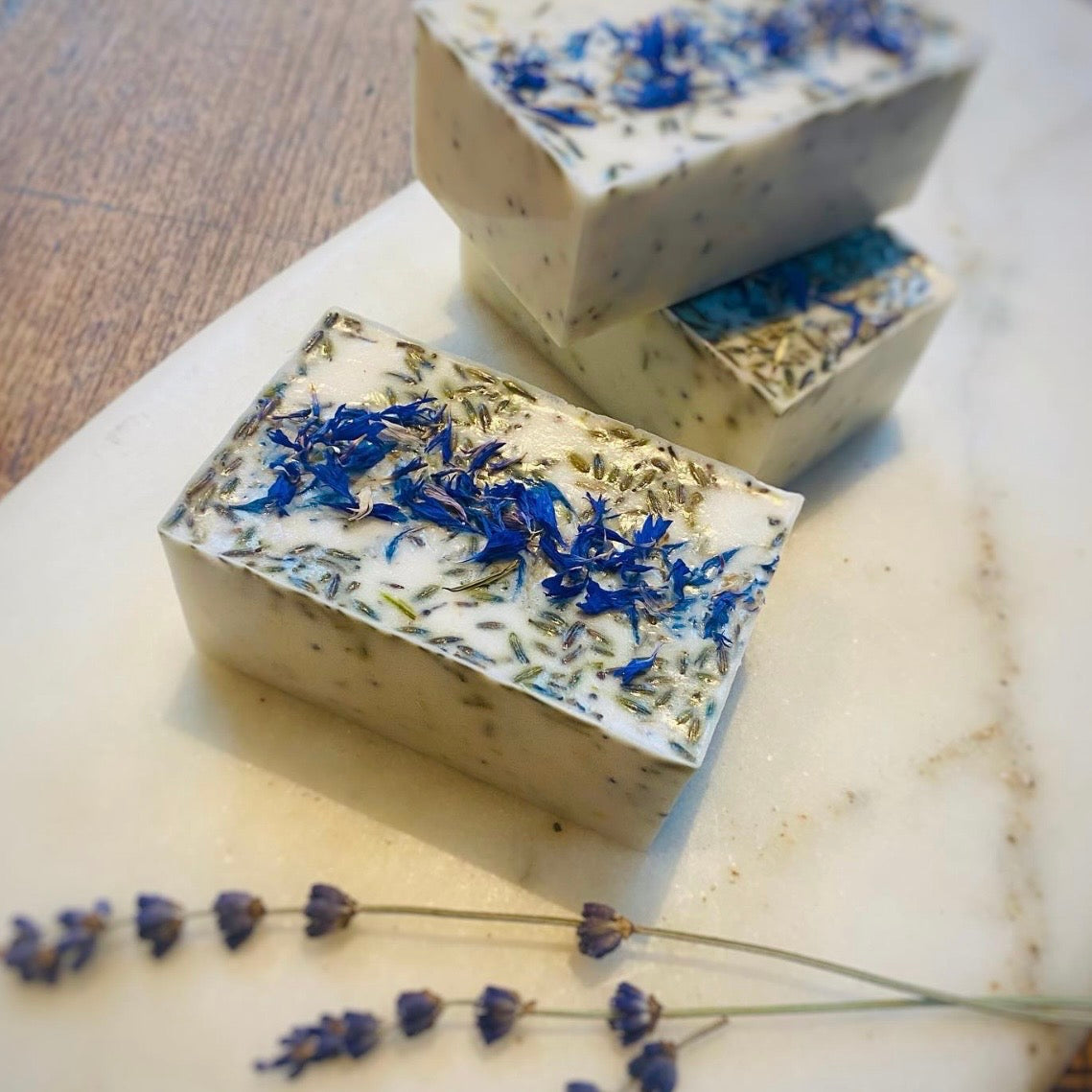 Three bars of soap with blue flower petals on a marble surface with lavender.