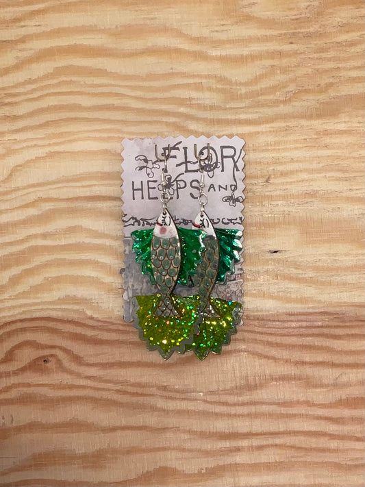 Green glittery earrings on a wooden surface with a visible brand label.