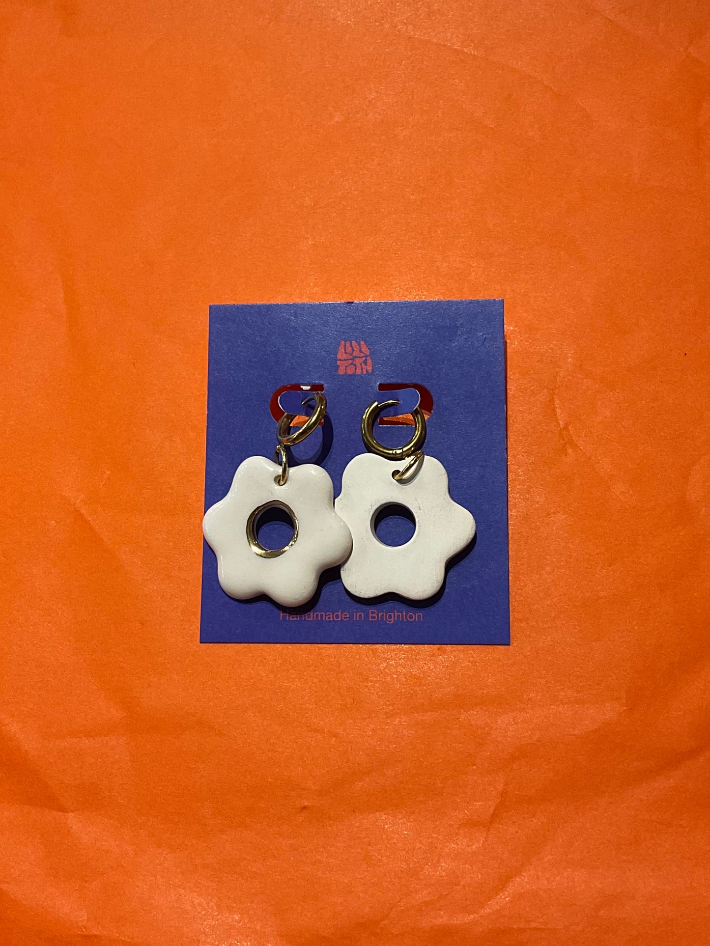 White flower-shaped earrings on a blue card with a visible brand logo against an orange background