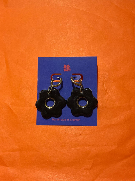 Black flower-shaped earrings on a blue card with 'Handmade in Belgium' text, against an orange background.