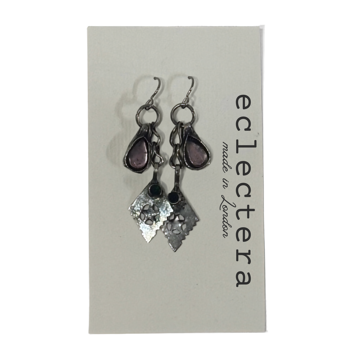 A pair of handmade dangle earrings displayed on a light gray brand card. The earrings feature silver-toned metal hooks and charms, including a teardrop-shaped purple stone and a textured hammered diamond-shaped silver charm.