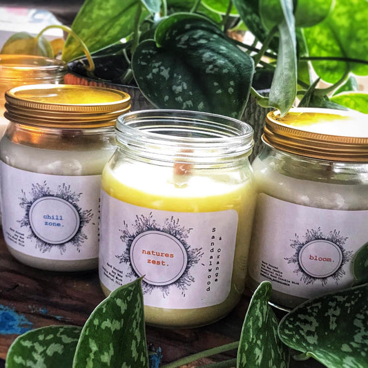 Three candles in glass jars with labels, surrounded by green plants.