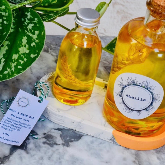 Two bottles of 'abeille' body and bath oil on a marble surface with a plant in the background.