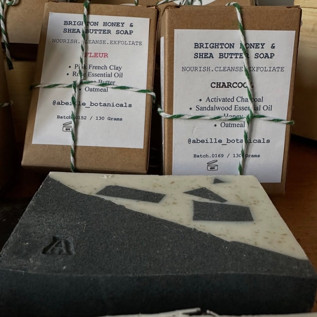 Two boxes of Brighton Honey & Shea Butter Soap with labels, one featuring French Clay and the other Charcoal, on a wooden surface.