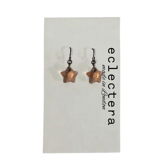 A pair of star-shaped drop earrings with dark metal hooks and a warm copper or rose-gold toned center, displayed on a white card printed with the brand name 'eclectera made in London'.