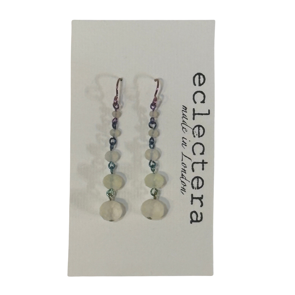 A pair of long beaded drop earrings displayed on a white card. The earrings feature a mix of translucent white beads, small dark beads, and silver-toned metal links and hooks.