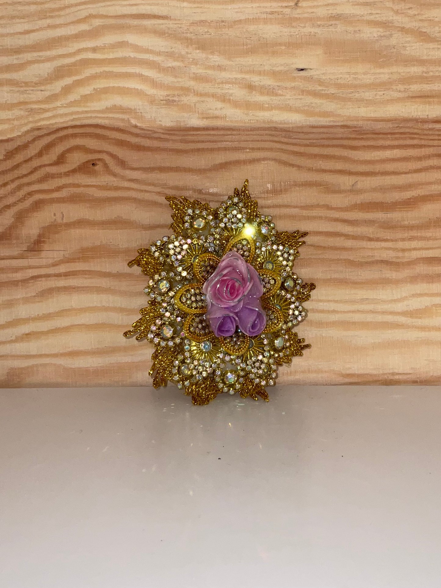 Decorative brooch with gold detailing and a pink and purple flower on a wooden surface.