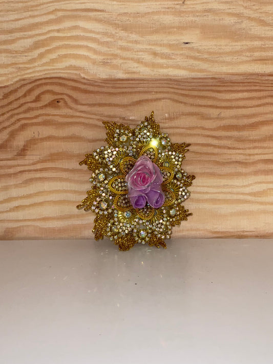 Decorative brooch with gold detailing and a pink and purple flower on a wooden surface.