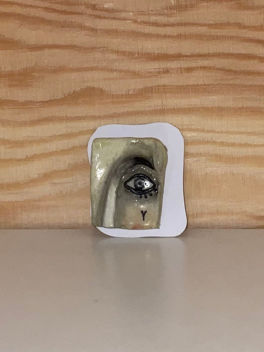 Small ceramic piece with a face design on a white card, against a wooden background