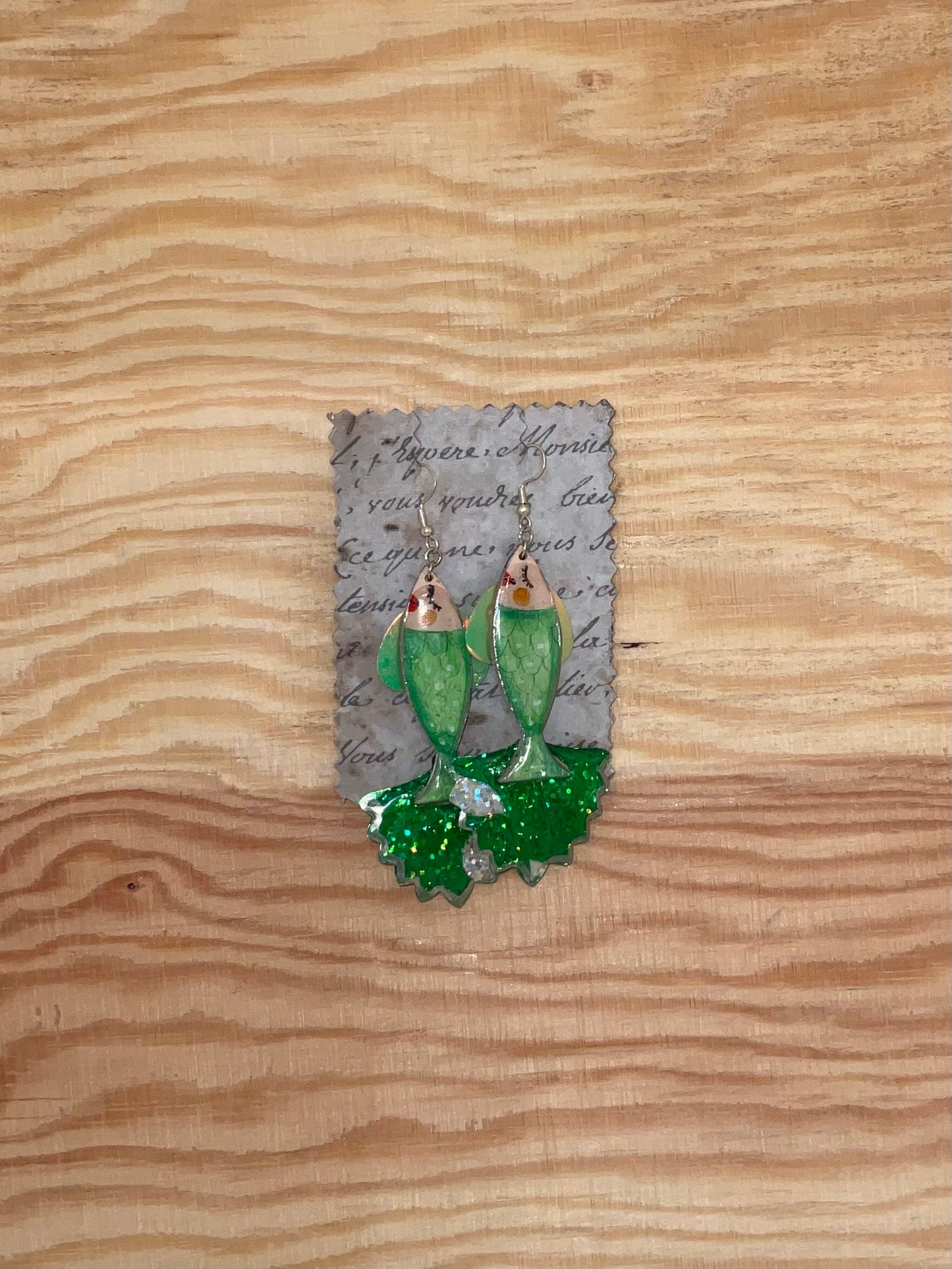 Mermaid-shaped earrings on a decorative card with text, placed on a wooden surface.