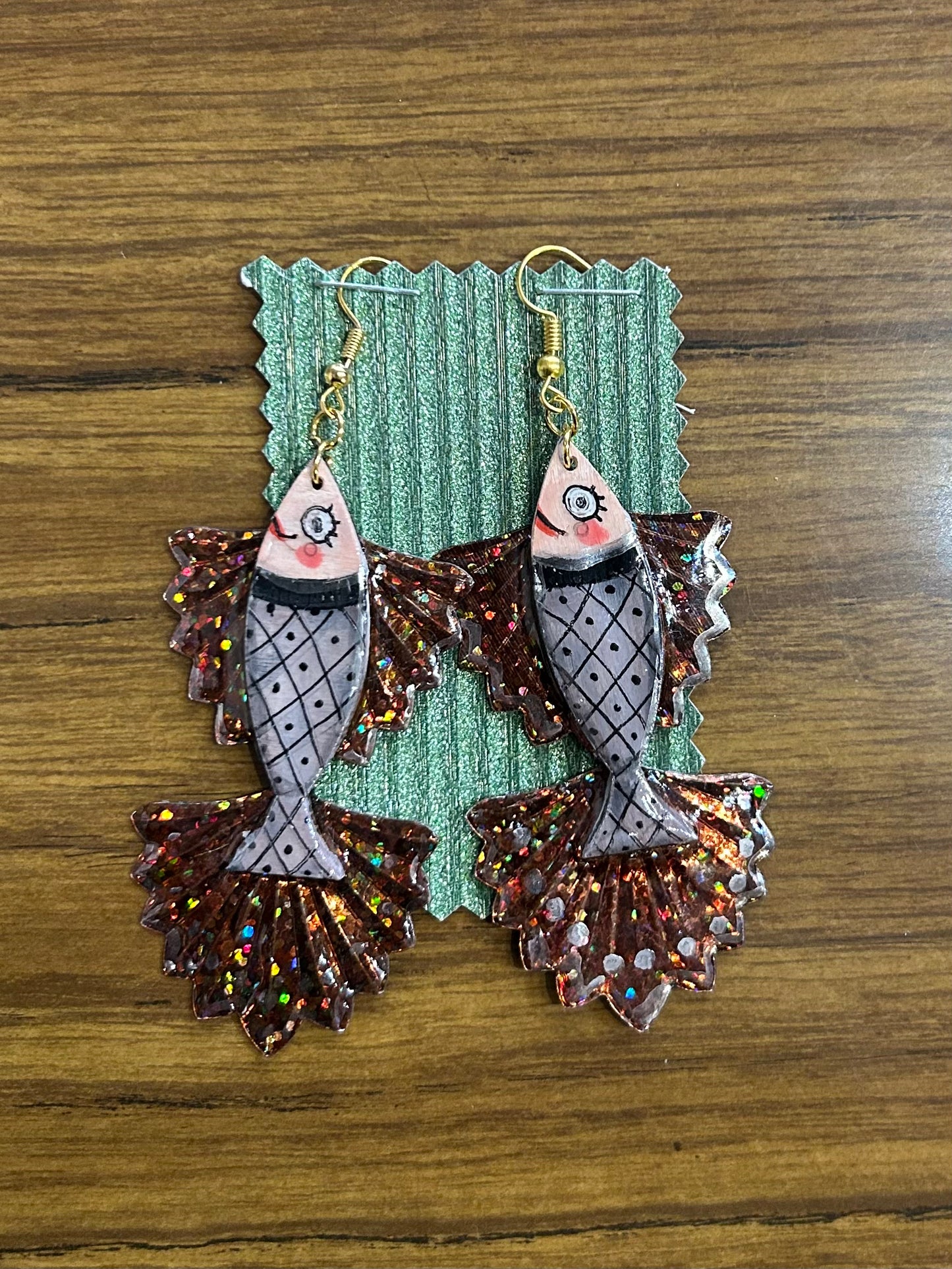 Pair of fish-shaped earrings with glittery details on a green card against a wooden background.