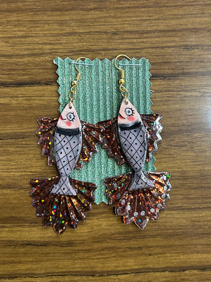 Pair of fish-shaped earrings with glittery details on a green card against a wooden background.
