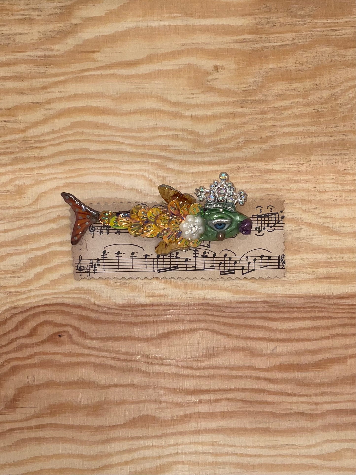Decorative fish brooch with gemstones on a piece of sheet music, placed on a wooden surface.