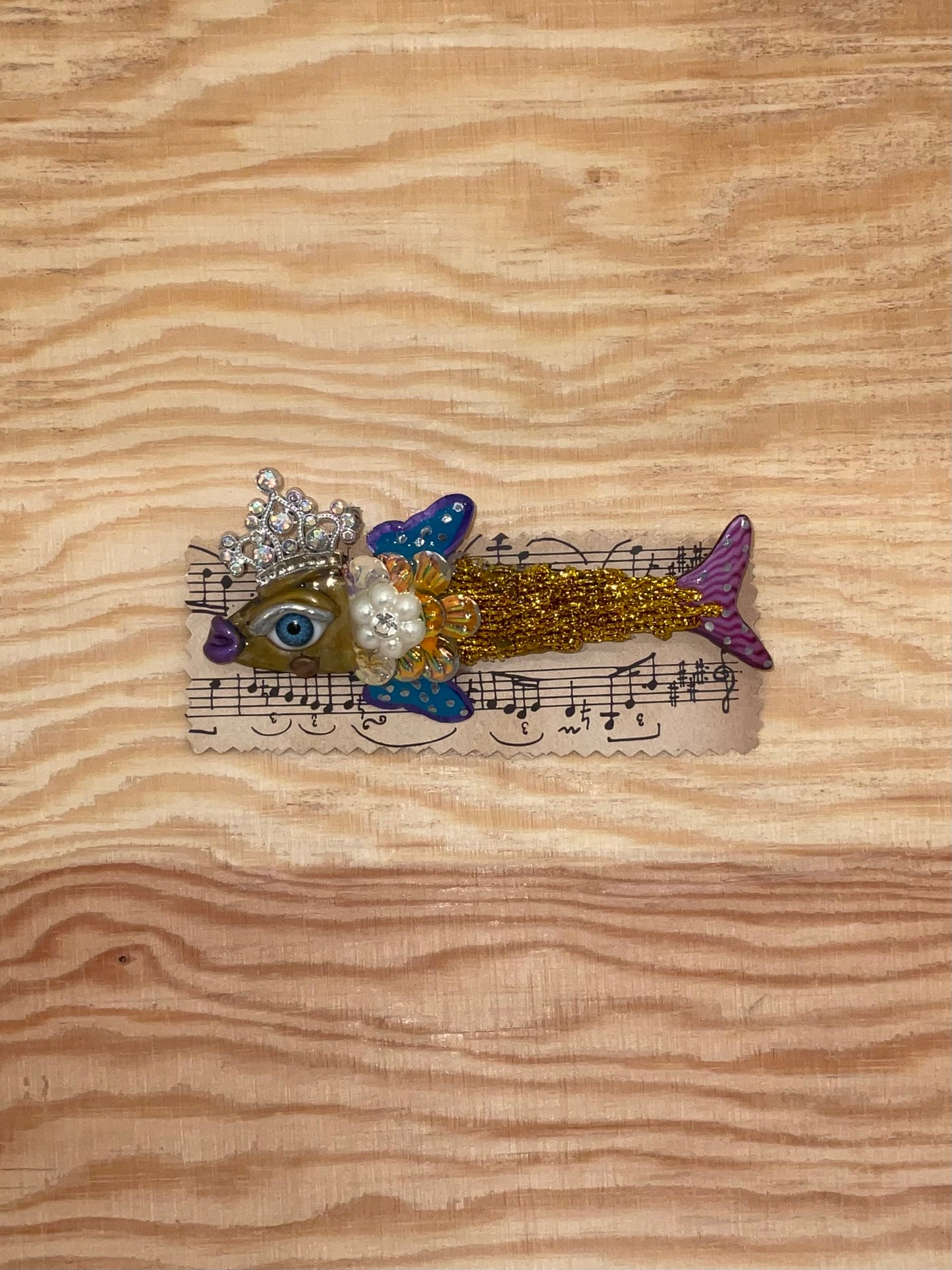 Decorative fish brooch with a crown on a piece of music sheet paper, placed on a wooden surface.
