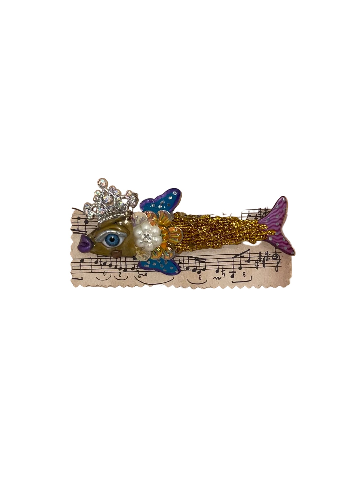 Decorative fish brooch with a crown on a piece of music sheet paper, placed on a wooden surface.