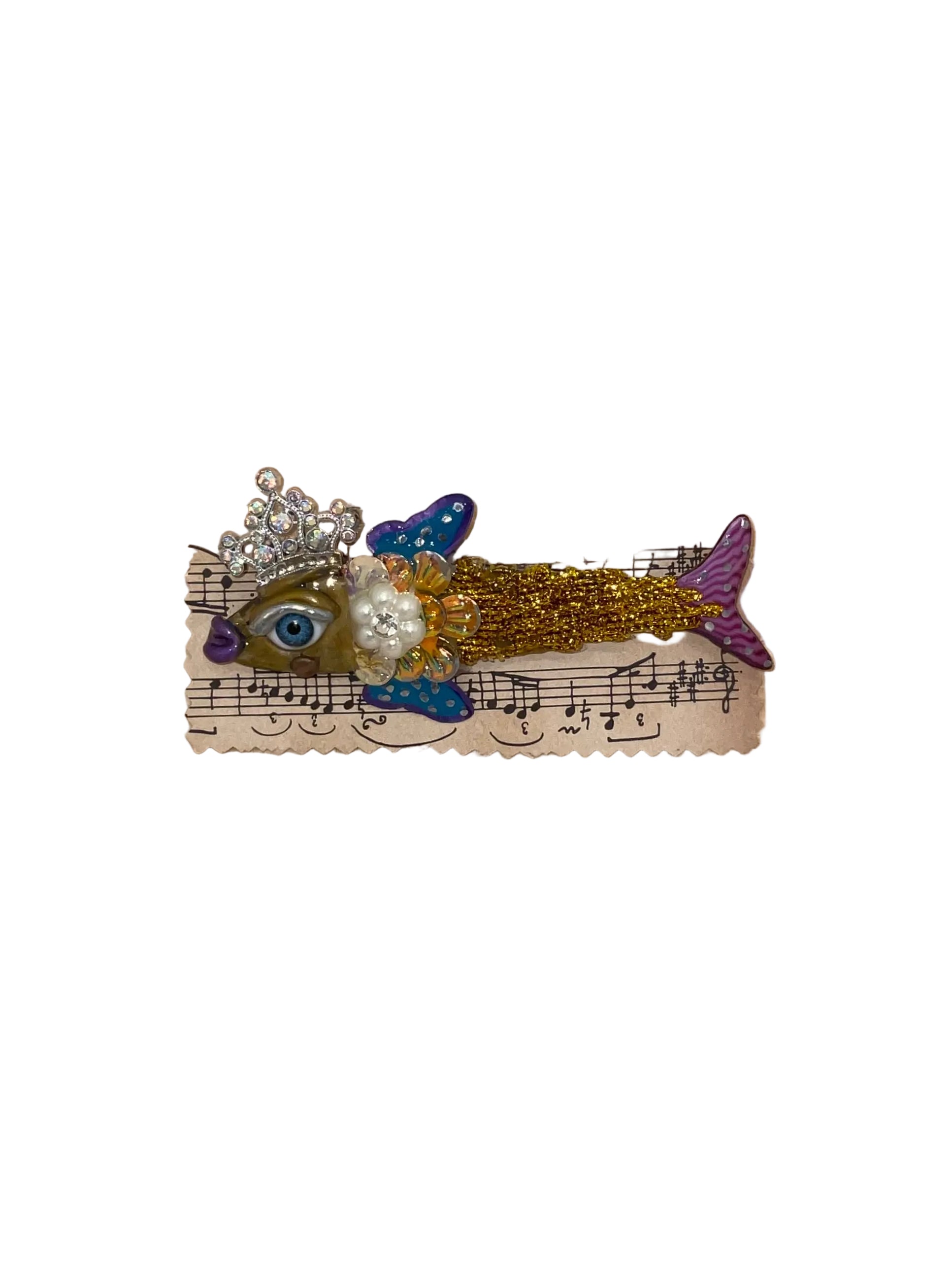Decorative fish brooch with a crown on a piece of music sheet paper, placed on a wooden surface.