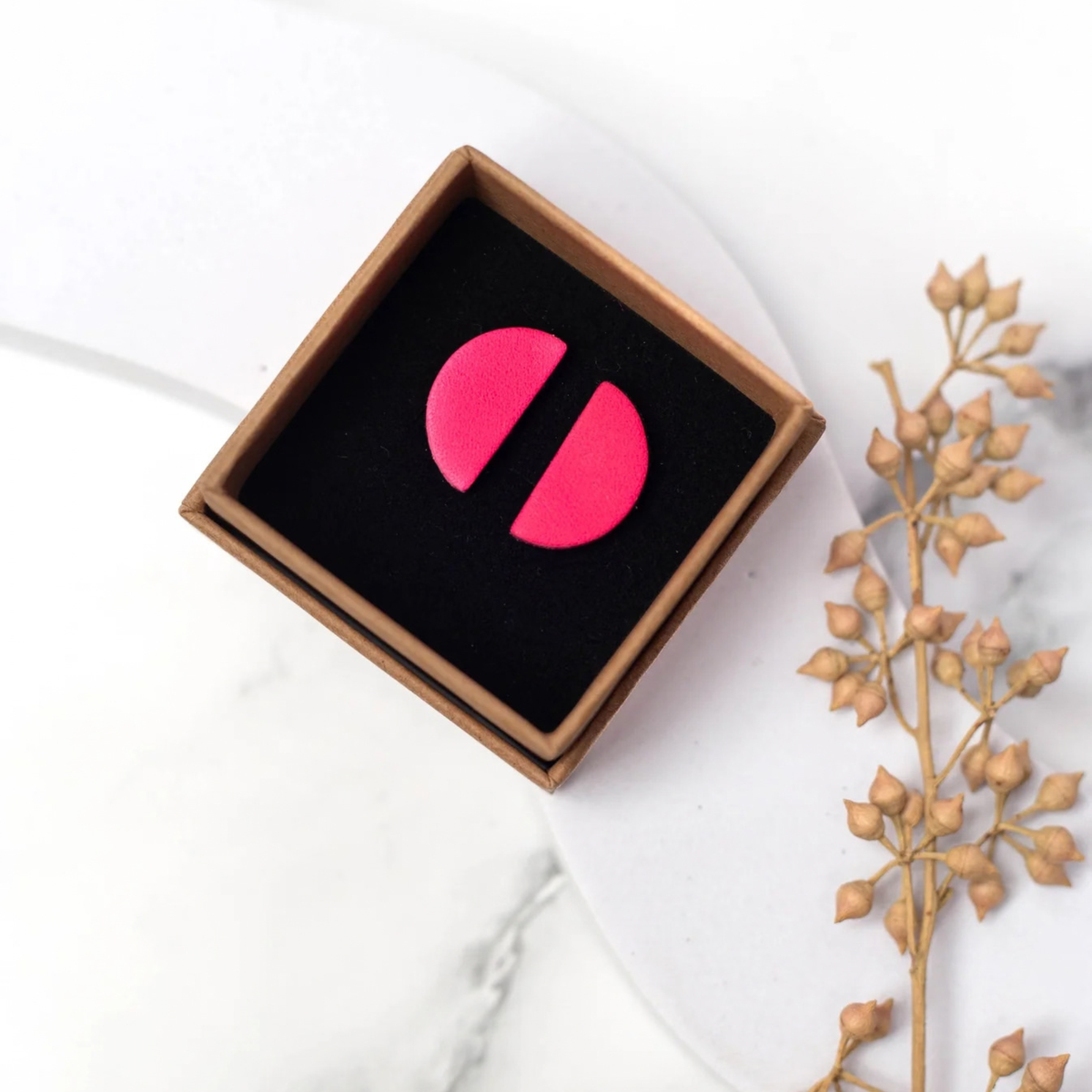 A pair of bright pink circular earrings displayed in a square brown gift box with a black velvet insert, set on a white marble surface next to a dried floral stem.