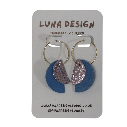A pair of gold hoop earrings with blue and glittery silver crescent moon drop charms, displayed on a grey LUNA DESIGN brand card.