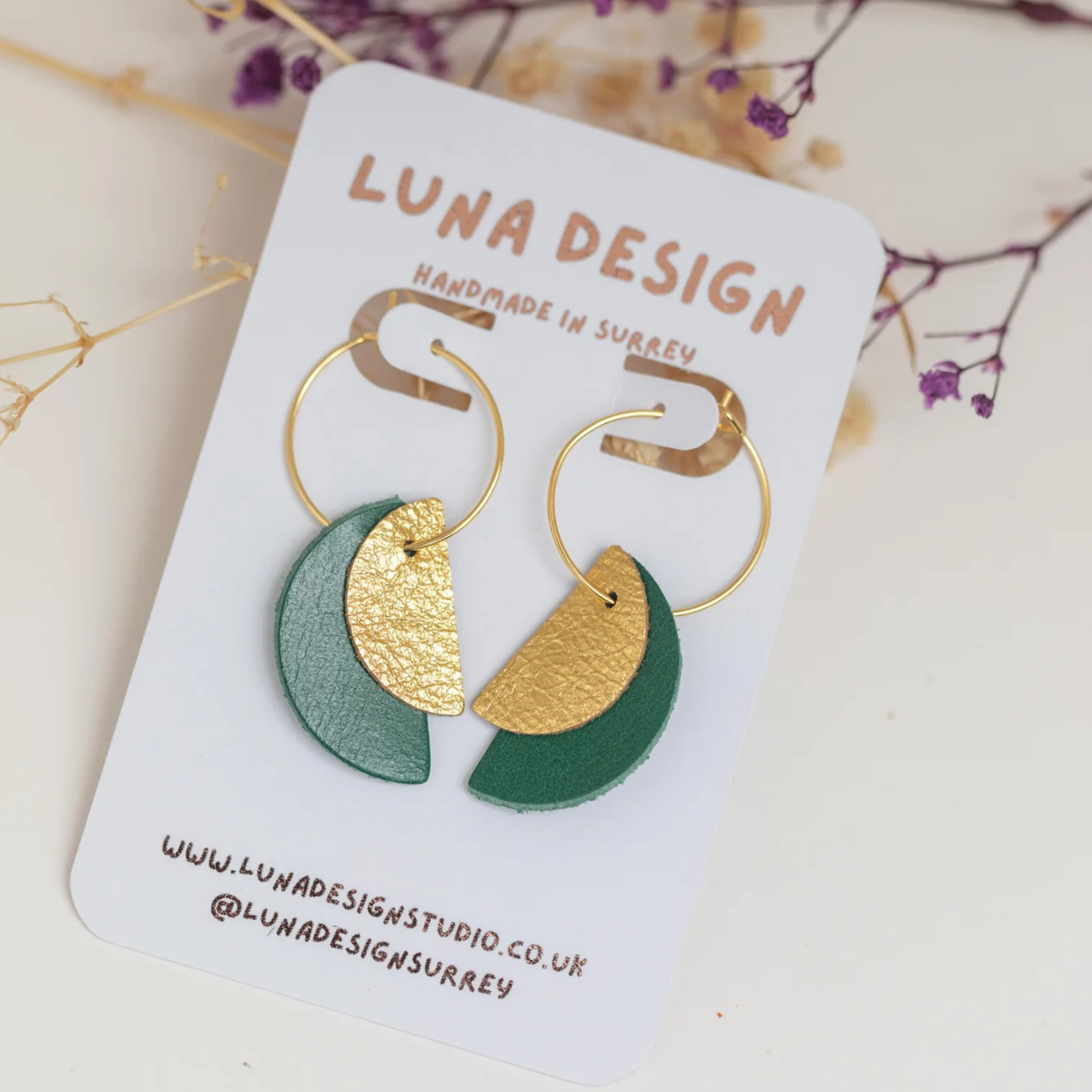 A pair of gold-tone hoop earrings with crescent moon and half-moon leather charms in dark green and textured gold, displayed on a white card labeled LUNA DESIGN.