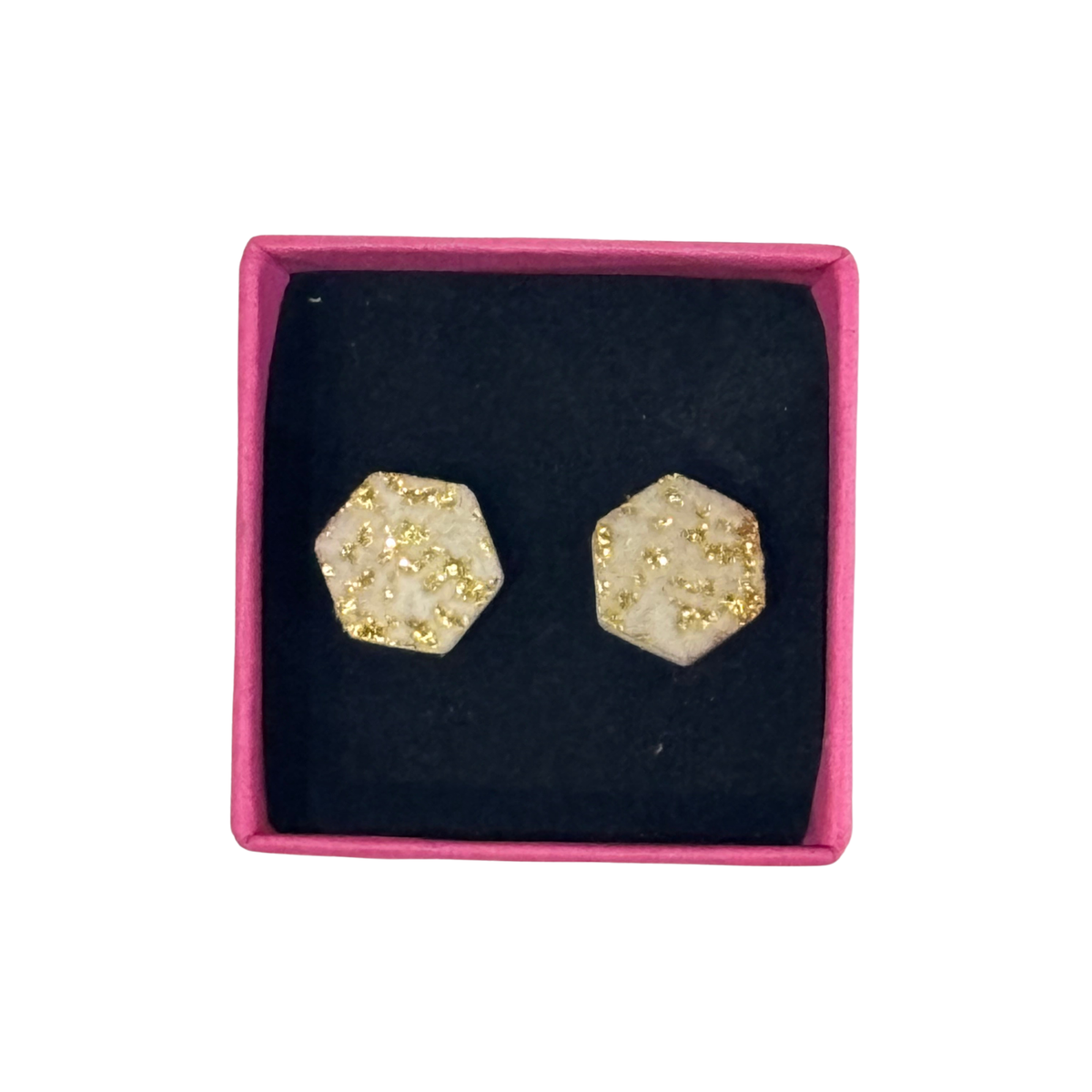 A pair of hexagonal stud earrings with a textured beige resin base and embedded gold glitter, presented in a pink box with a black velvet insert.