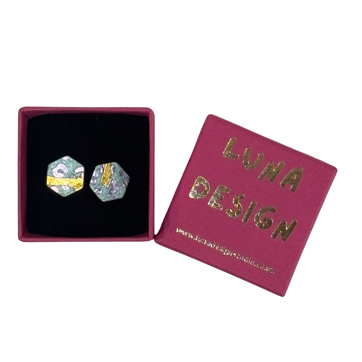 A pair of hexagonal stud earrings displayed in an open magenta jewelry box next to its lid, which features gold foil lettering that reads 'LUNA DESIGN'. The earrings have a multicolor abstract pattern with green, blue, yellow, and white tones.