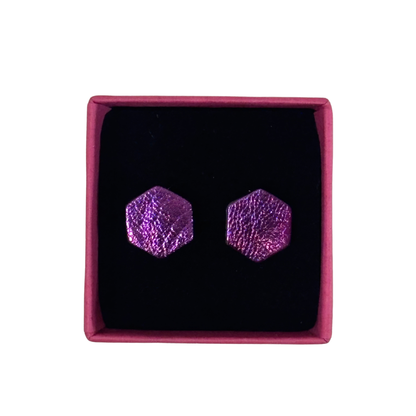 A pair of textured hexagonal stud earrings in a vibrant purple metallic finish, presented inside a square magenta box with a black velvet insert, set against a plain white background.