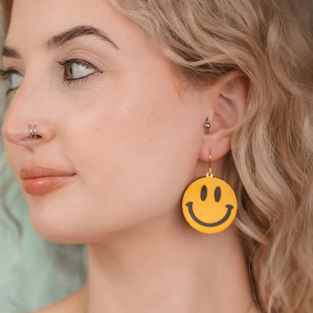 A close-up of a woman wearing a yellow smiley face dangle earring. The earring features a round yellow face with black eyes and a smile, hanging from a gold-tone hook.