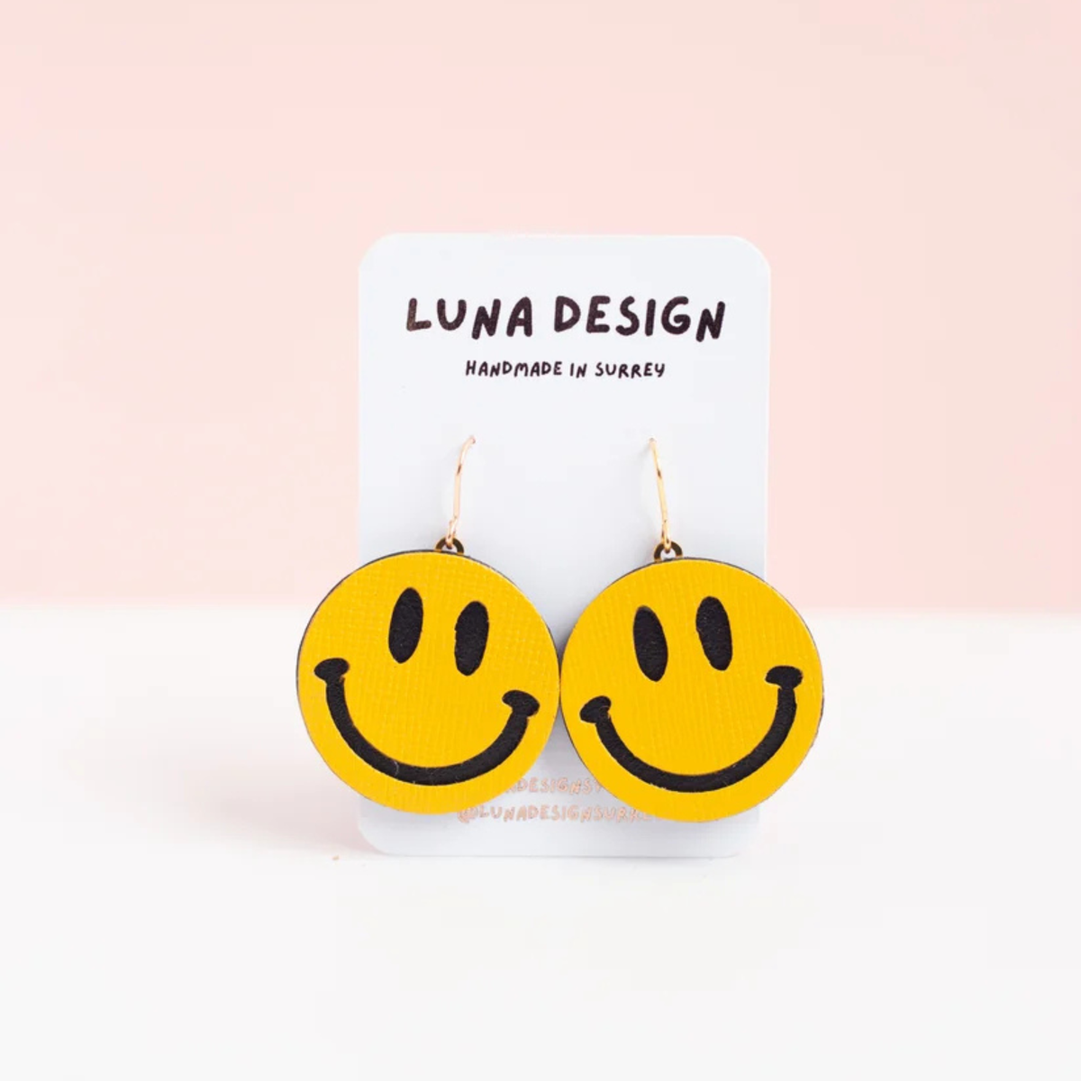 A pair of yellow smiley face drop earrings with black outlines, displayed on a white card labeled 'LUNA DESIGN' against a soft pink and white background.