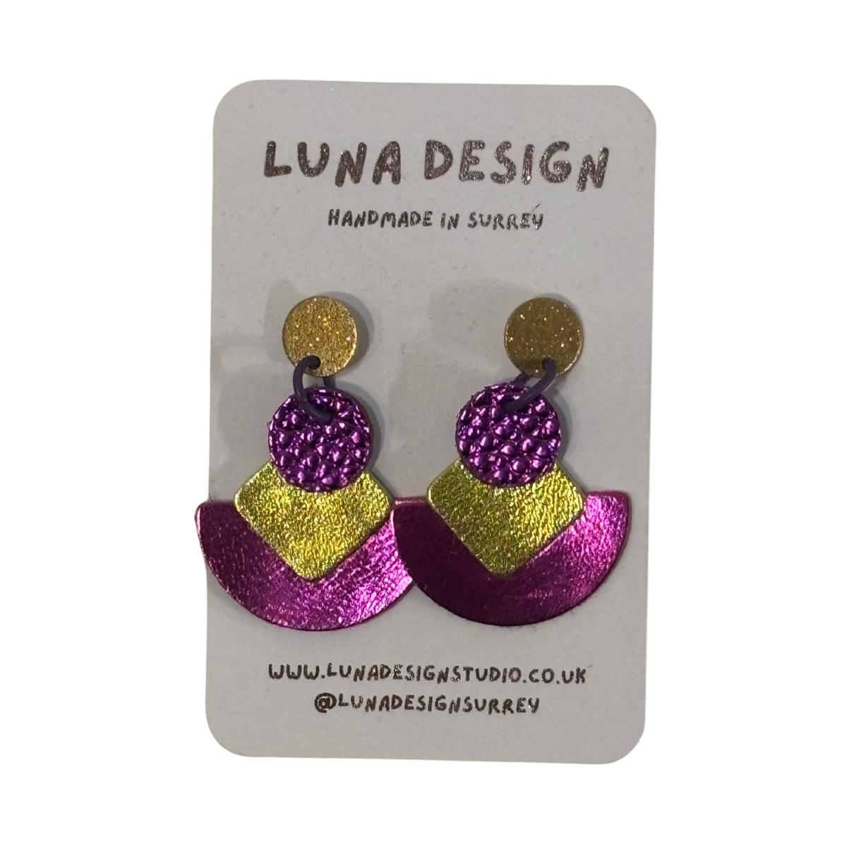A pair of handmade statement earrings by Luna Design, featuring a gold glitter stud, a textured purple glitter circle, and a split purple and gold glitter chevron, displayed on a white card backing.