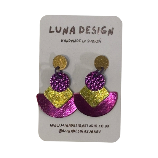 A pair of handmade statement earrings by Luna Design, featuring a gold glitter stud, a textured purple glitter circle, and a split purple and gold glitter chevron, displayed on a white card backing.