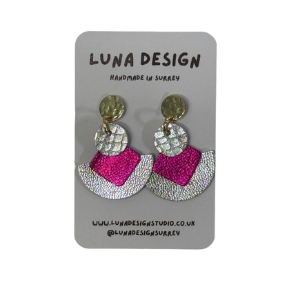 A pair of handmade statement earrings by Luna Design, featuring textured gold-tone studs, silver-tone circular discs, and fan-shaped drops in hot pink and silver glitter, displayed on a grey card backing.