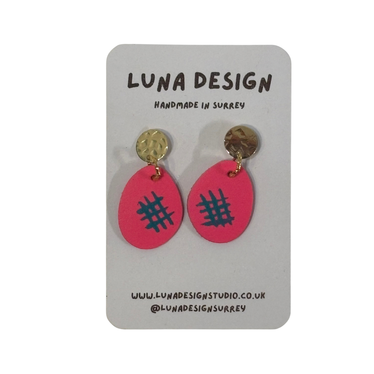 A pair of handmade dangle earrings by Luna Design, featuring bright pink oval drops with a black grid pattern and textured gold circular studs, displayed on a grey card backing.