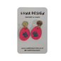 A pair of handmade dangle earrings by Luna Design, featuring bright pink oval drops with a black grid pattern and textured gold circular studs, displayed on a grey card backing.