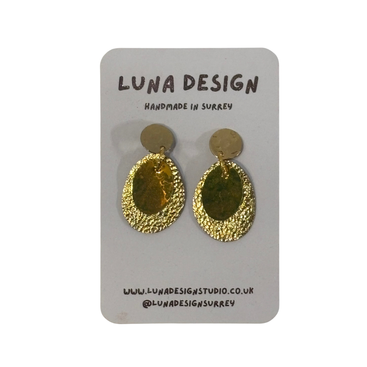 A pair of textured gold teardrop-shaped dangle earrings with flat circular gold studs, displayed on a grey card backing printed with 'LUNA DESIGN'.