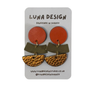 A pair of handmade geometric dangle earrings by Luna Design, featuring a terracotta orange disc, a textured olive green chevron, and a textured gold half-moon, displayed on a white branded card.