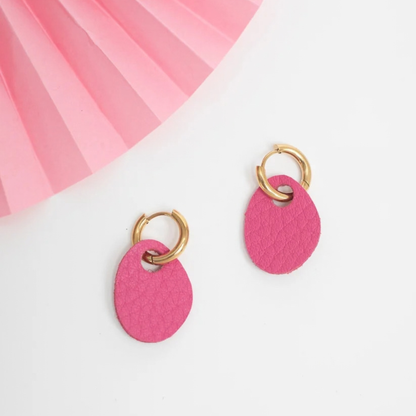A pair of gold hoop earrings with textured hot pink oval drop charms, displayed on a clean white surface with a pink pleated paper fan in the top-left corner.