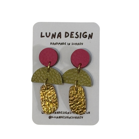 A pair of handmade dangle earrings by Luna Design, featuring a magenta pink circle, a textured olive green semicircle, and a hammered gold textured drop, displayed on a white backing card.