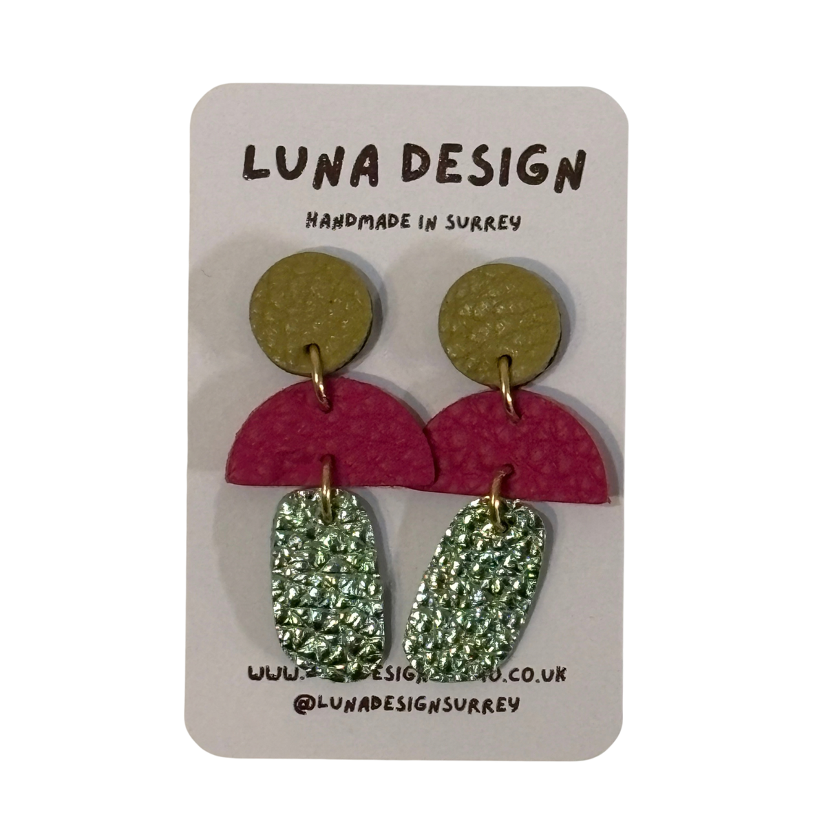 A pair of handmade drop earrings by Luna Design, featuring olive green textured discs, magenta semicircles, and glittery green teardrop charms, displayed on a white card backing.