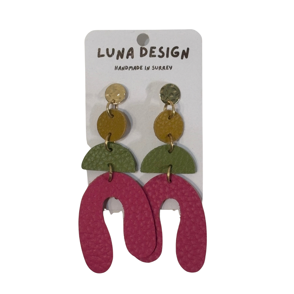 A pair of handmade statement earrings by Luna Design, featuring a gold-tone top stud with a textured gold disc, a green semicircle, and a large textured magenta U-shaped drop. The earrings are displayed on a white cardboard backing card that reads 'LUNA DESIGN HANDMADE IN SURREY'.