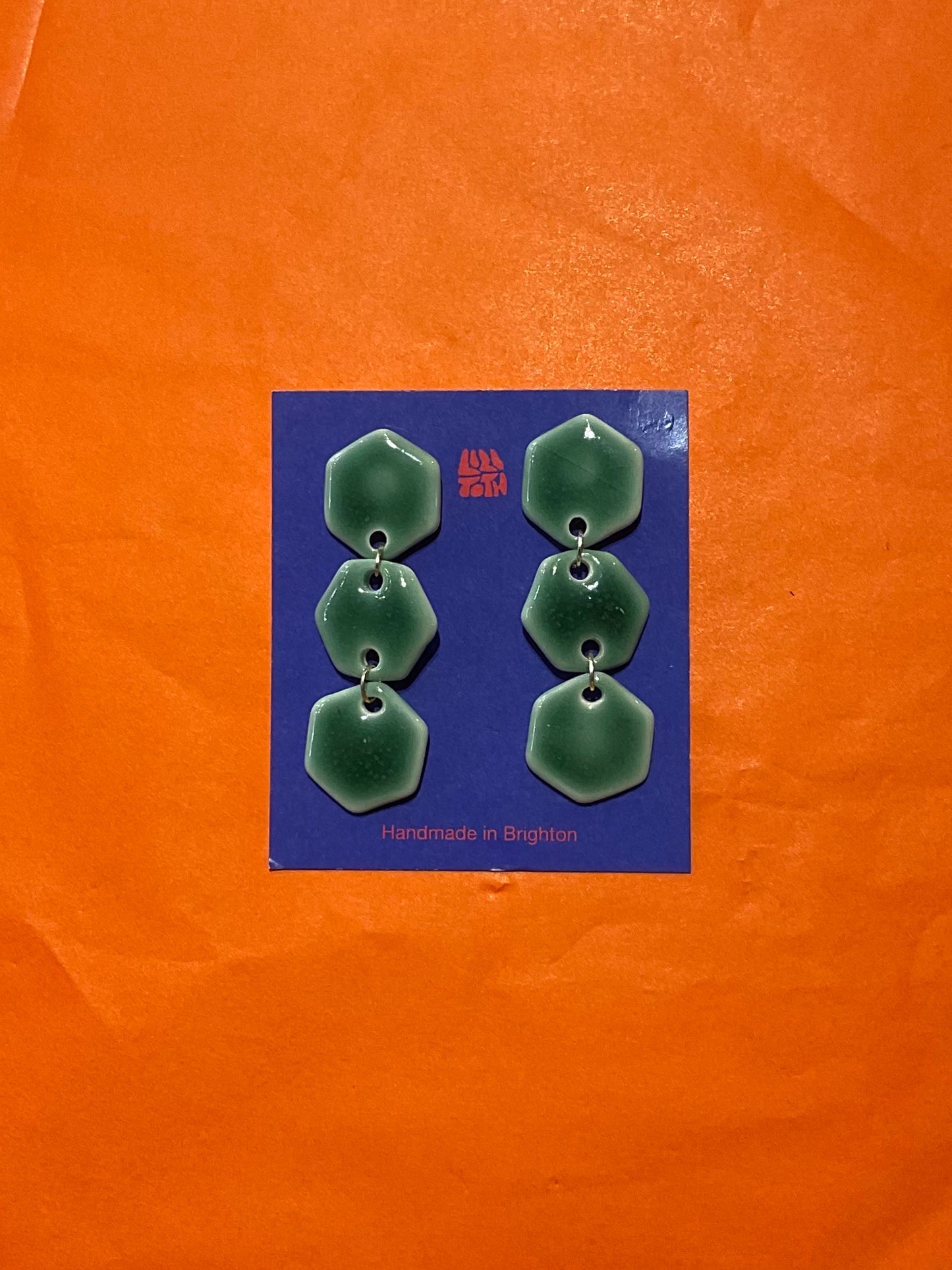 Green earrings on a blue card with 'Handmade in Brighton' text, against an orange background