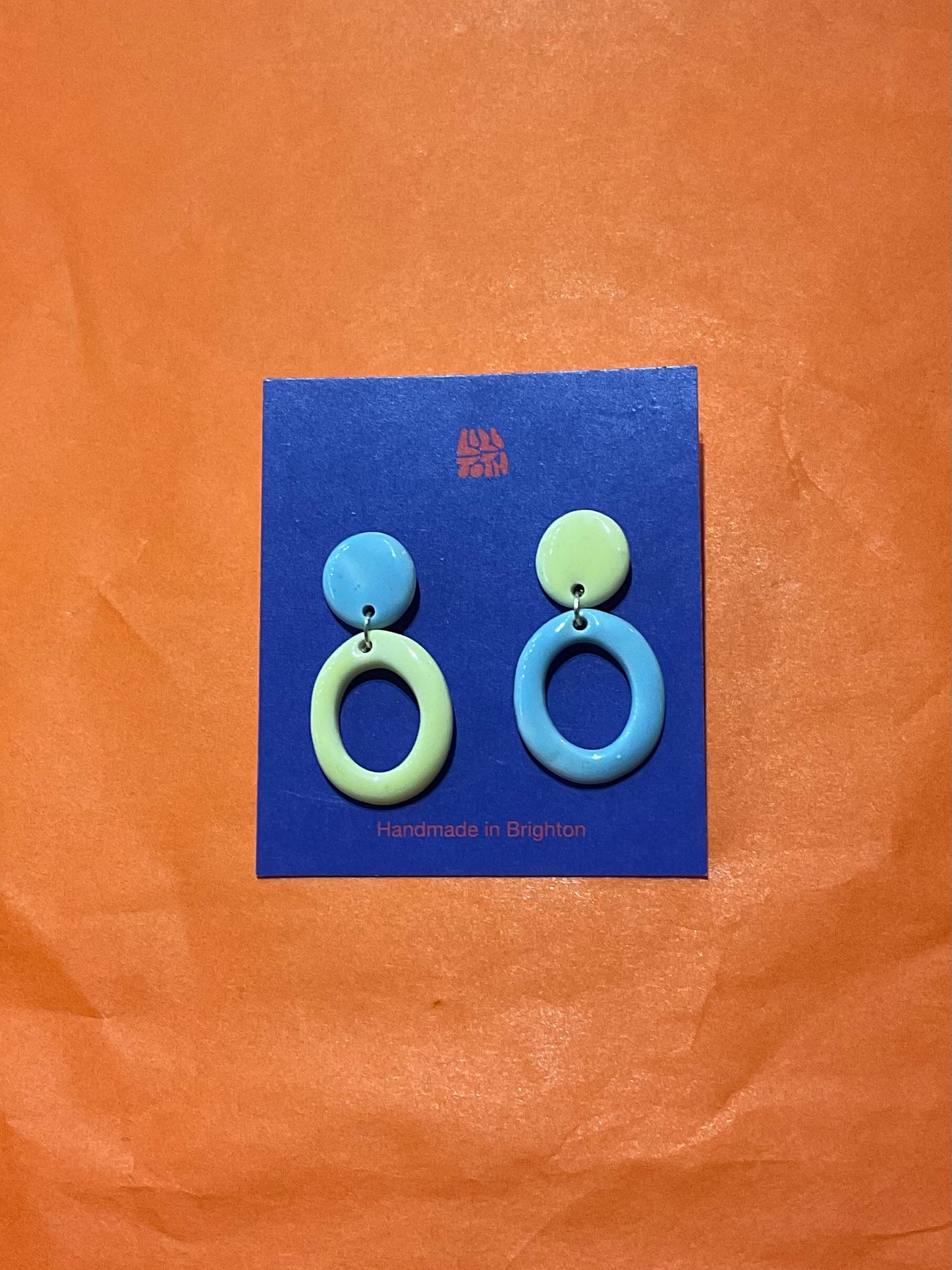 Pair of blue and green circular earrings on a blue card against an orange background