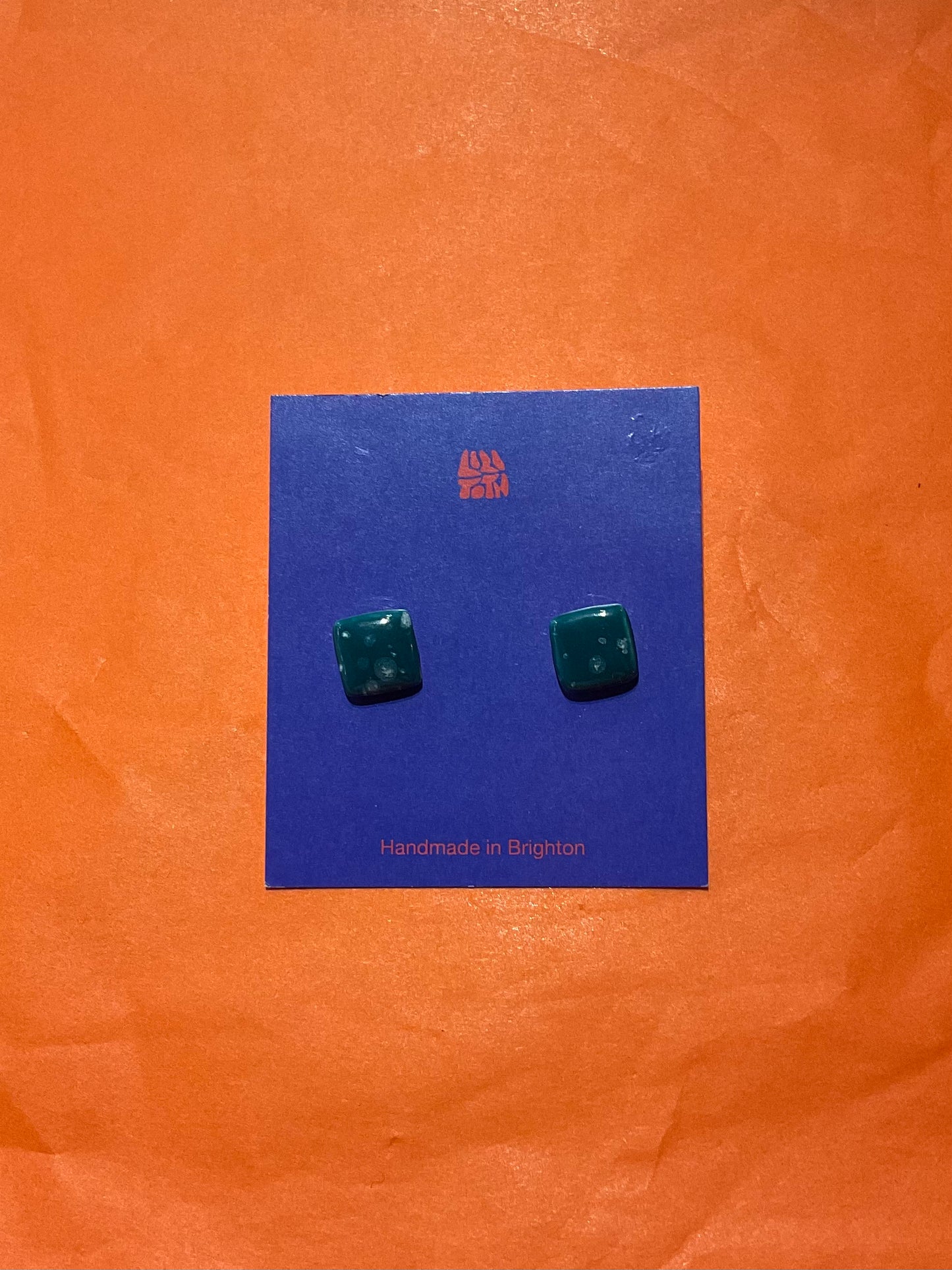 Green square earrings on a blue card with 'Handmade in Brighton' text, against an orange background.