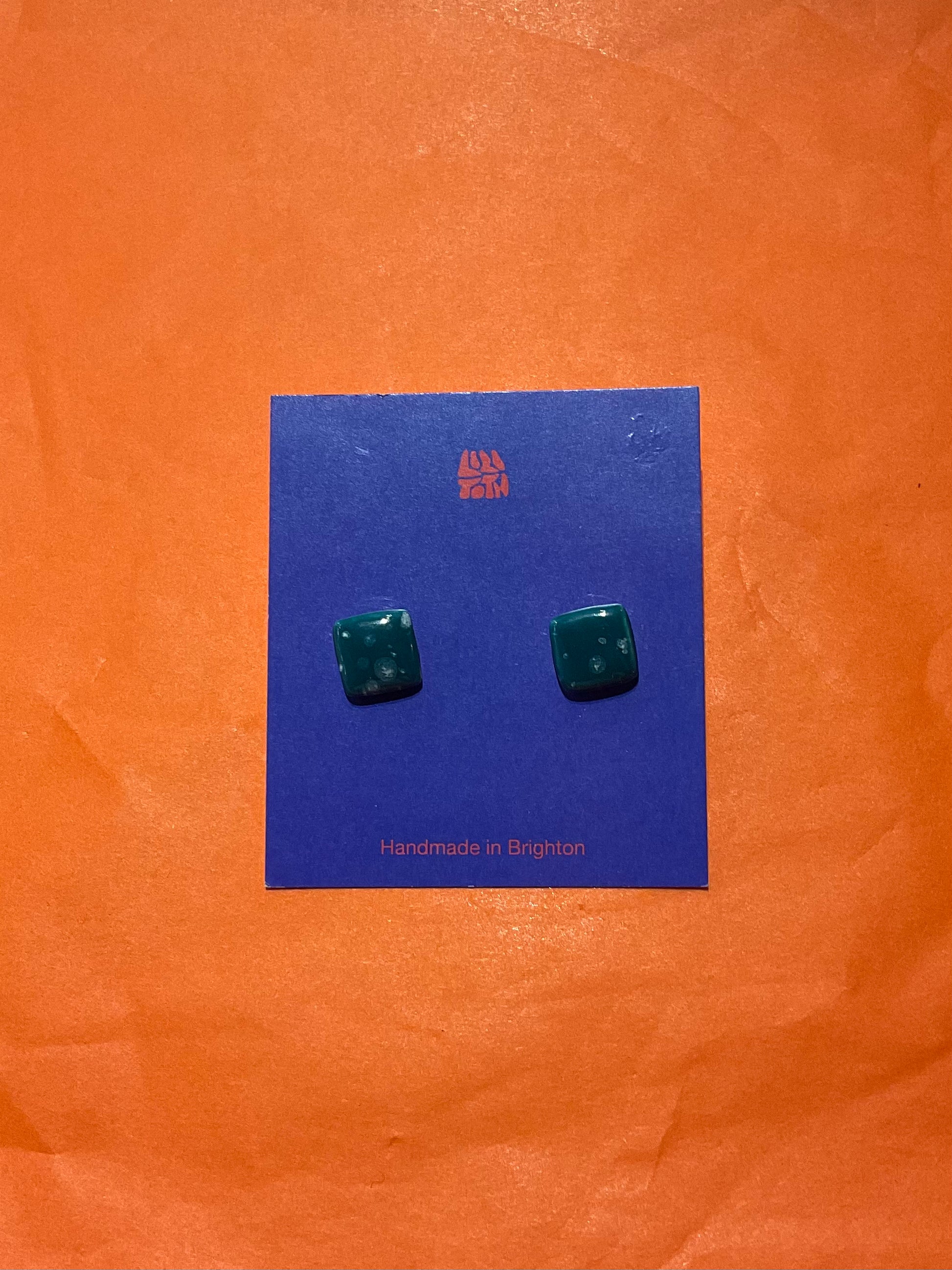 Green square earrings on a blue card with 'Handmade in Brighton' text, against an orange background.
