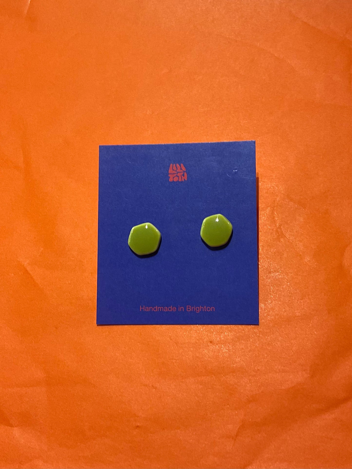 Green hexagonal earrings on a blue card with 'Handmade in England' text, against an orange background.
