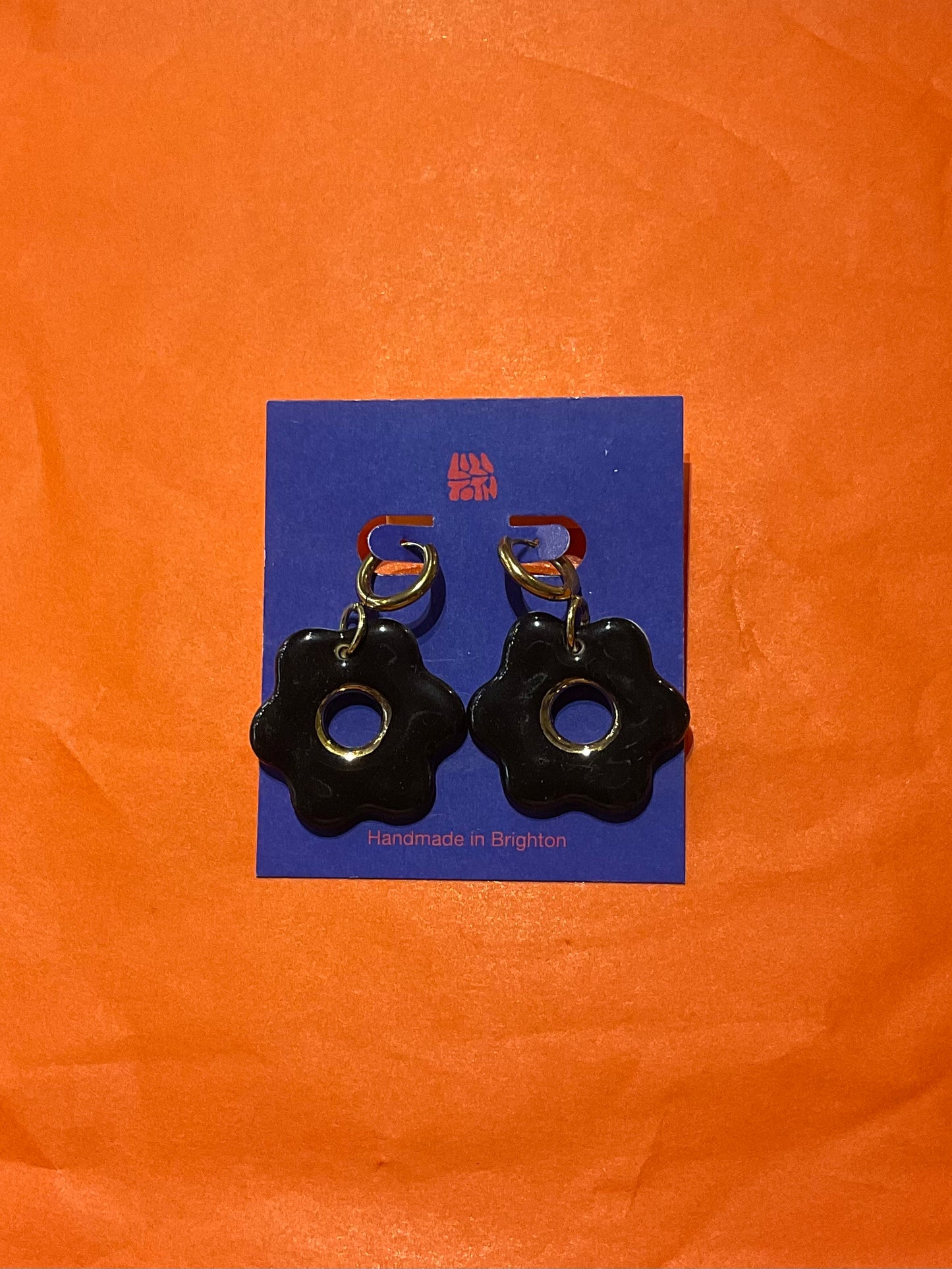 Black flower-shaped earrings on a blue card with 'Handmade in Belgium' text, against an orange background.