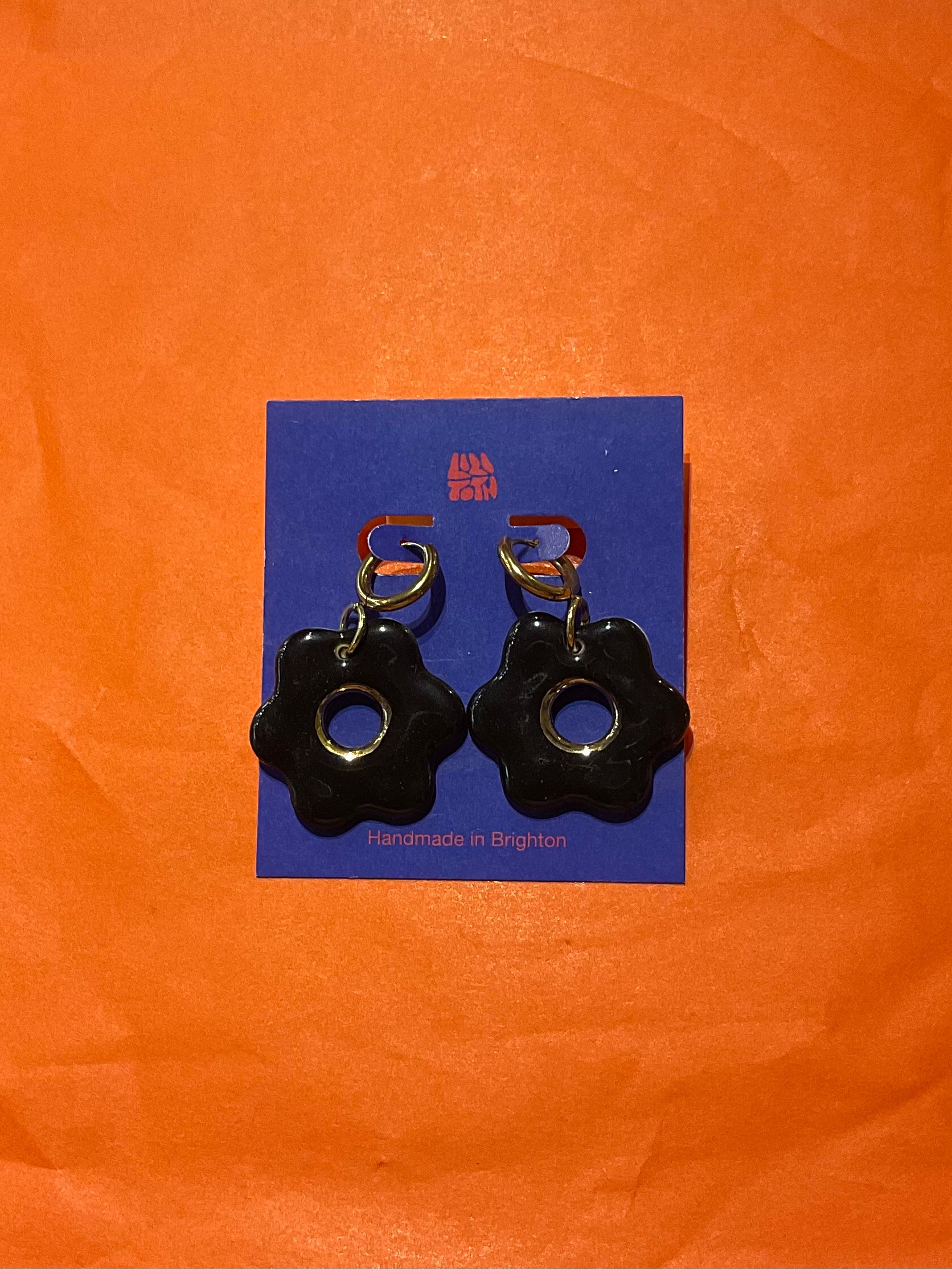 Black flower-shaped earrings on a blue card with 'Handmade in Belgium' text, against an orange background.