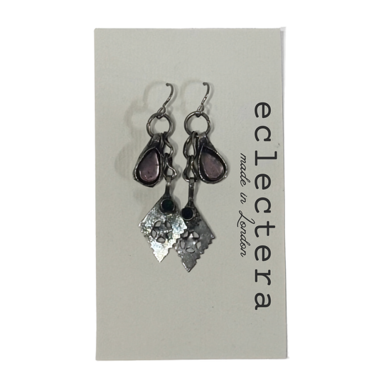 A pair of handmade dangle earrings displayed on a light gray brand card. The earrings feature silver-toned metal hooks and charms, including a teardrop-shaped purple stone and a textured hammered diamond-shaped silver charm.
