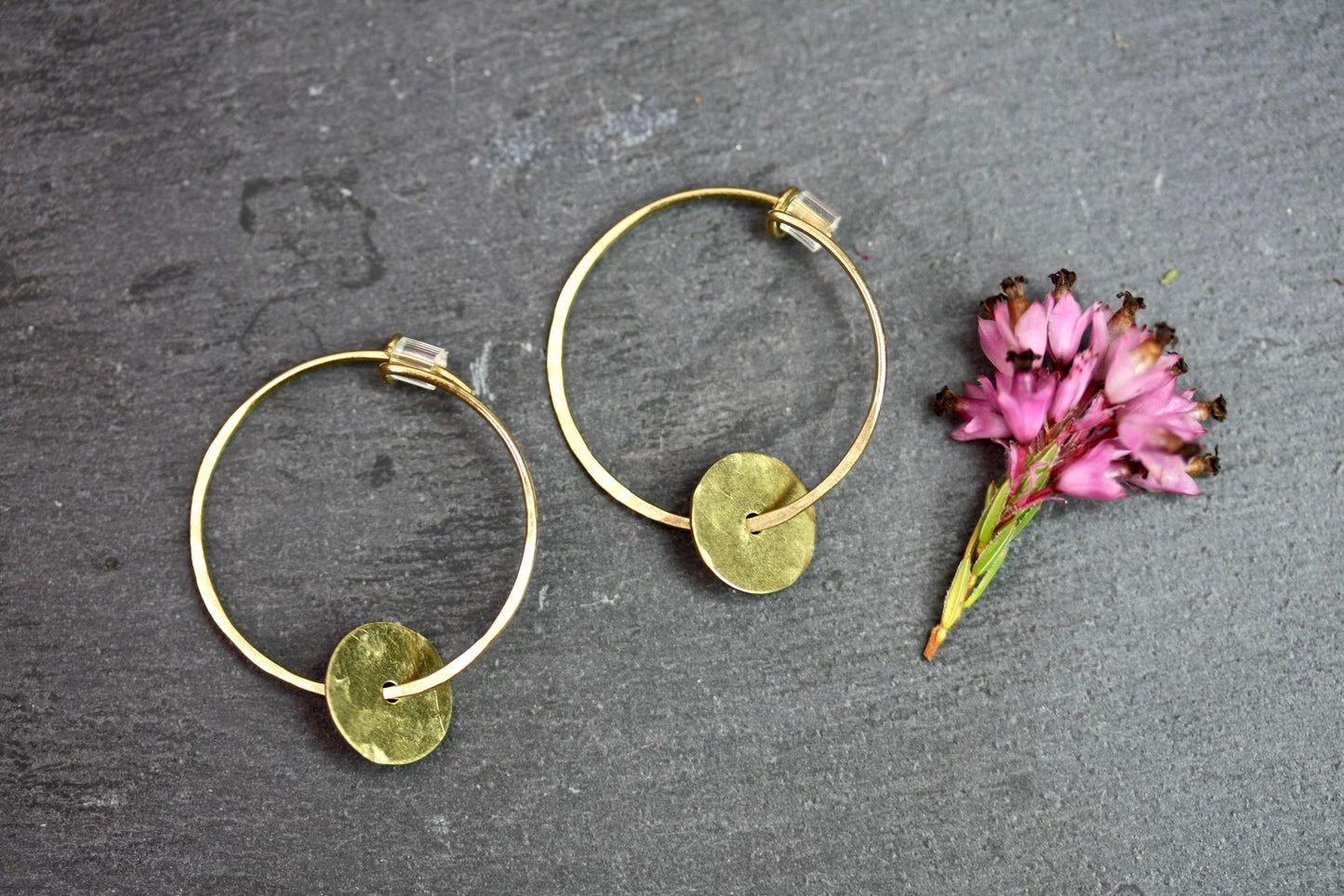 Two gold hoop earrings with circular discs on a dark surface with a small pink flower.
