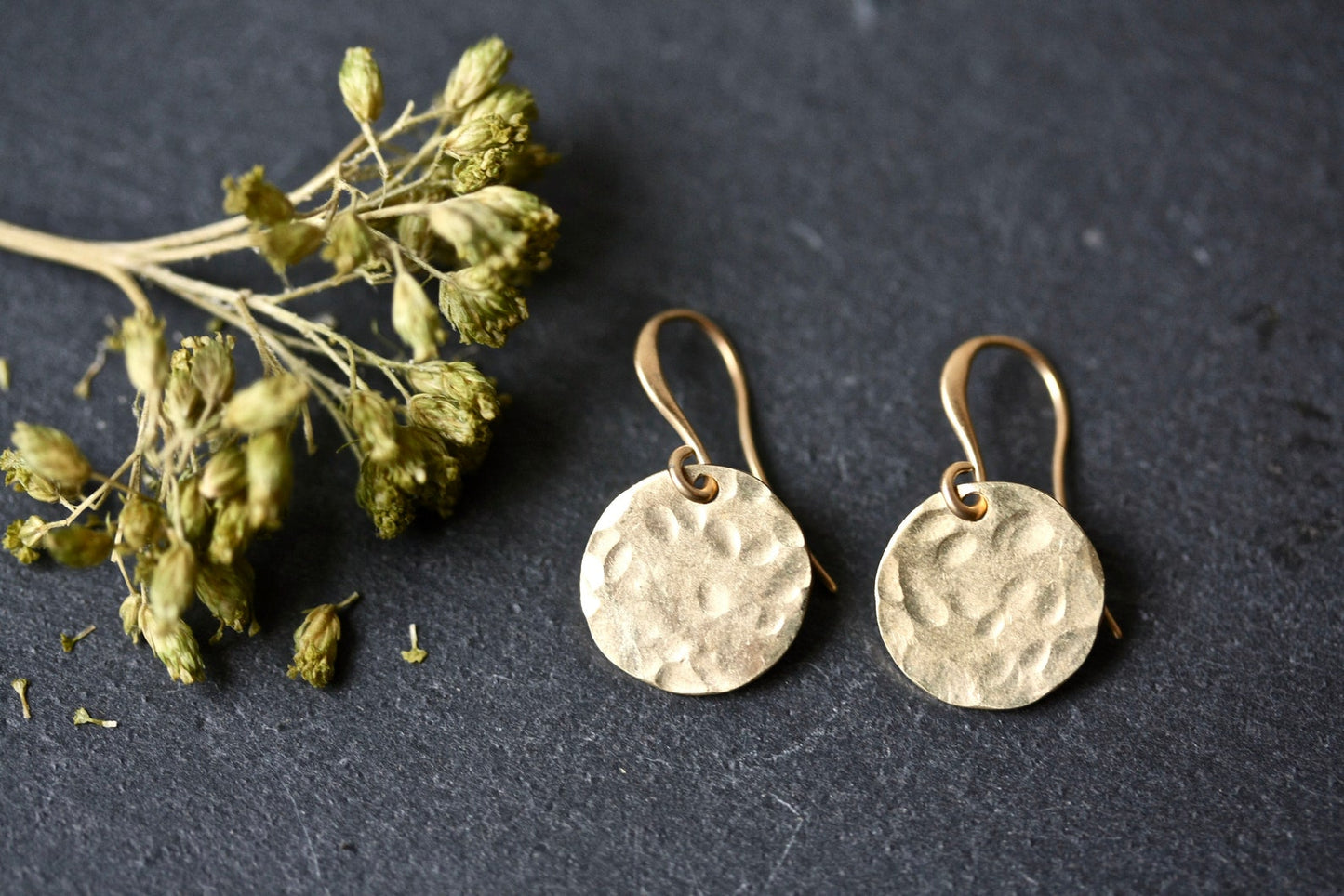 Pair of gold earrings on a dark surface with dried greenery.