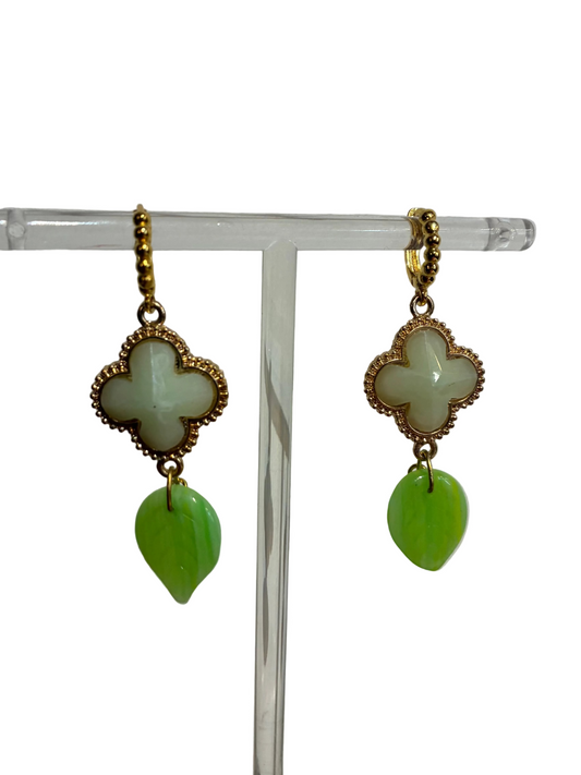 Pair of earrings with gold clover-shaped tops and green leaf-like bottoms on a clear stand against a white background.